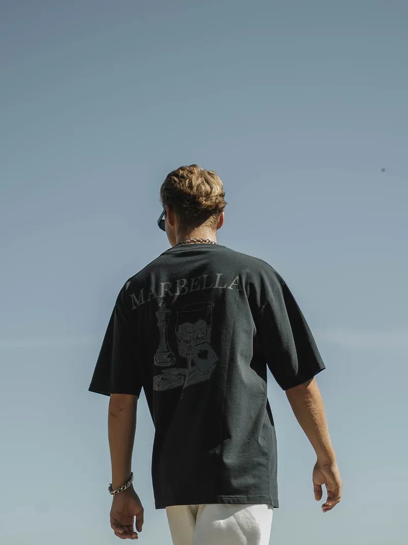 A black tee printed with Marbella, a chess king, a glass and cards, against the sky